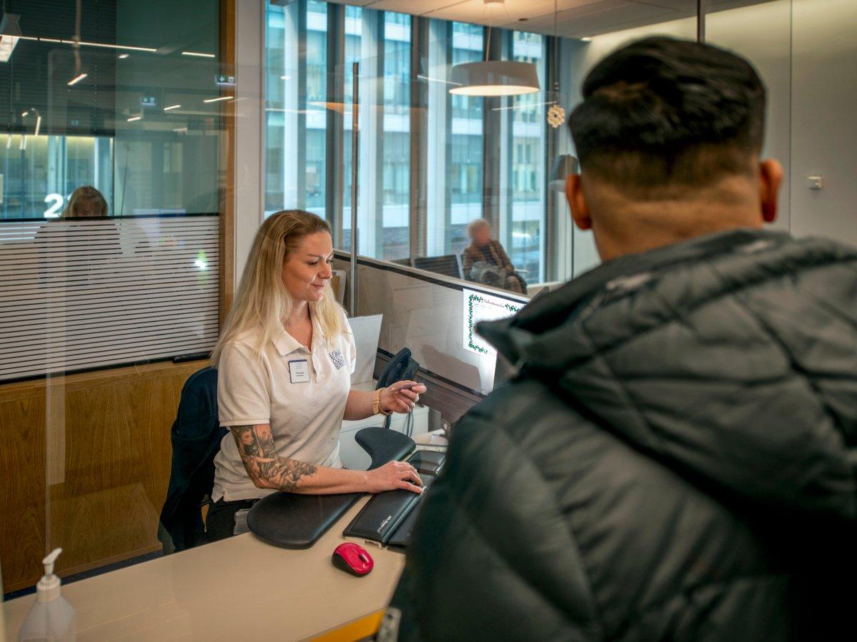 En kvinna i vit t-shirt sitter framför en dator. I förgrunden syns en patient i svart täckjacka.