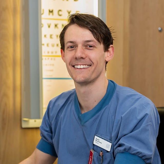 En man i sjukhuskläder sitter framför en dator. Bakom honom syns en syntavla.