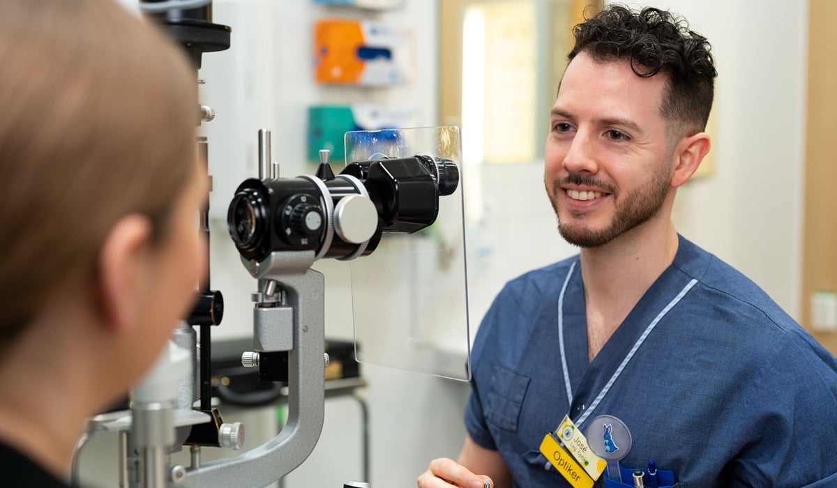 En man i sjukhuskläder sitter bredvid ett mikroskop och tittar mot en patient.