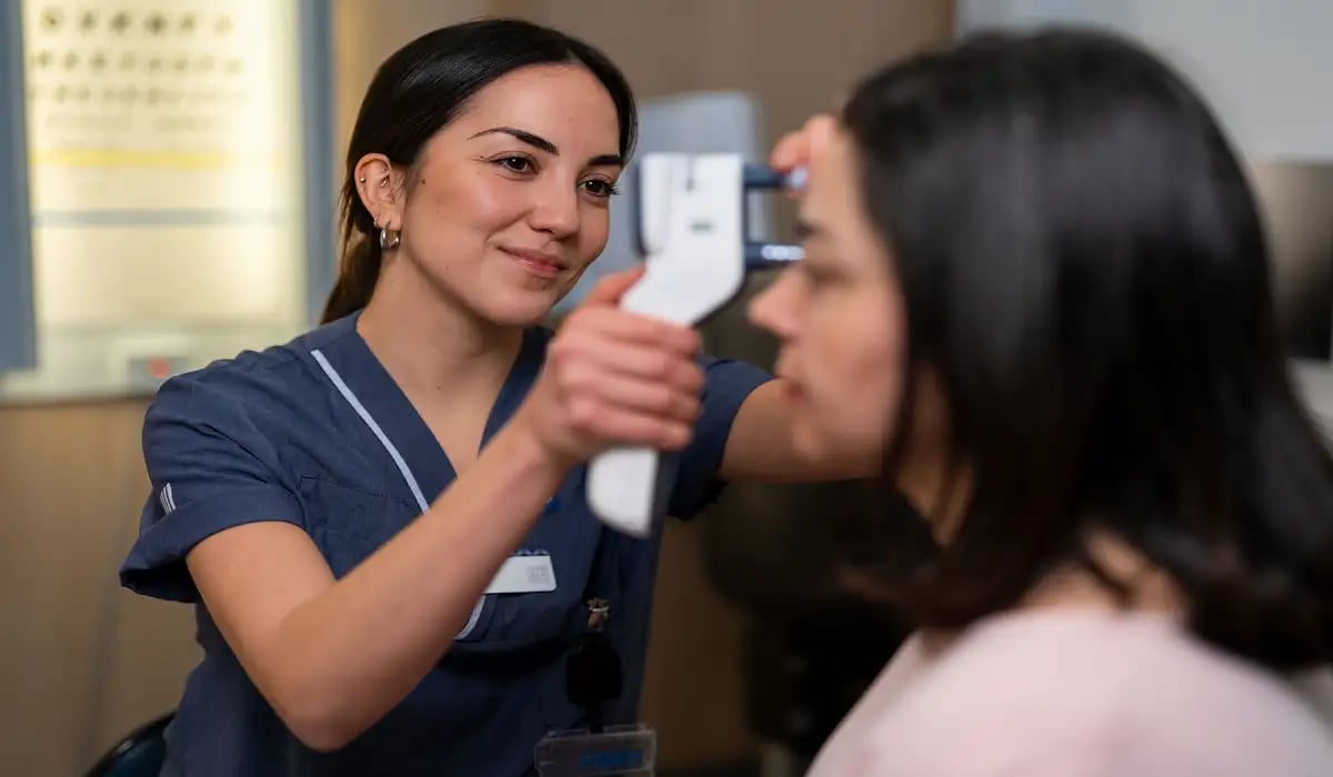 En person i sjukhuskläder undersöker ögat på en patient. Patienten sitter vänd åt sidan, medan vårdpersonalen ler vänligt och koncentrerat genomför undersökningen.