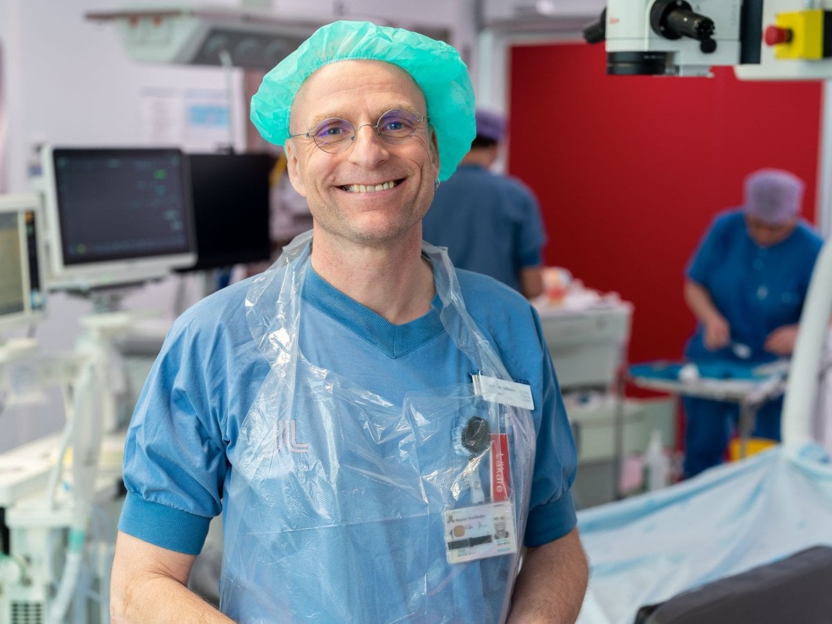 En person i blå arbetskläder, plastförkläde och grön skyddsmössa ler mot kameran i ett operationsrum. I bakgrunden syns medicinsk utrustning, monitorer och vårdpersonal