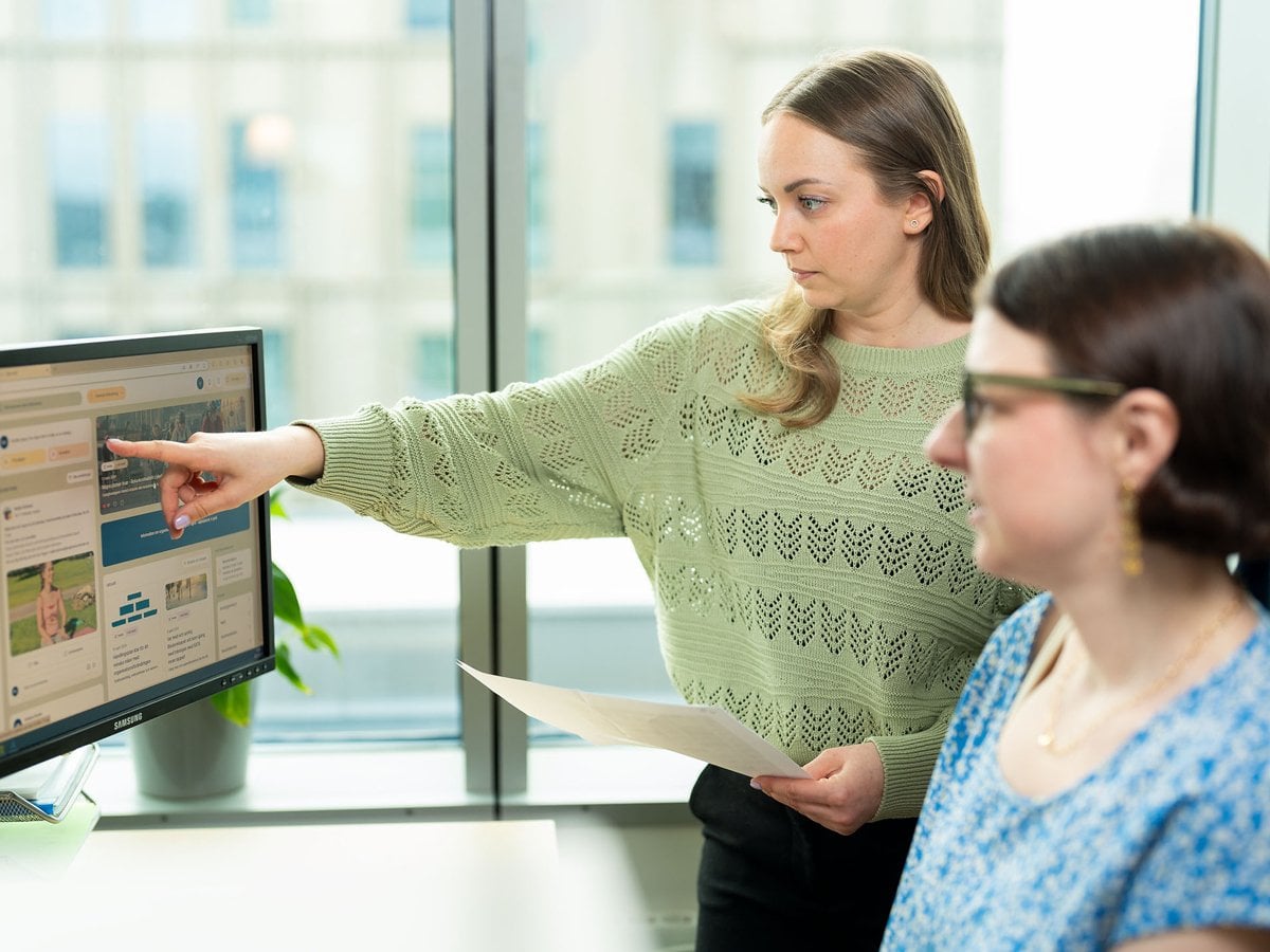 Två personer arbetar tillsammans vid en dator. Den ena personen, klädd i en grön tröja, pekar på något på datorskärmen medan den andra, klädd i en blåmönstrad topp, sitter och tittar på skärmen.