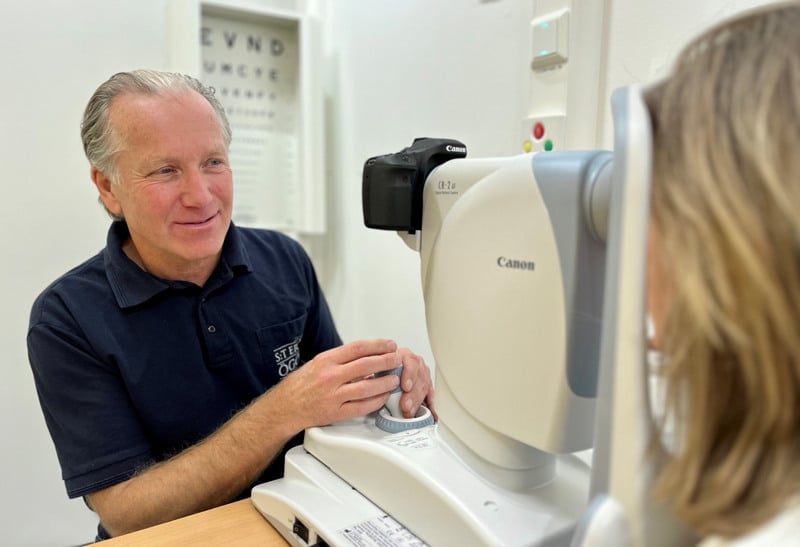 En man i blå pikétröja, sitter vid en ögonbottenkamera och tittar mot en patient.