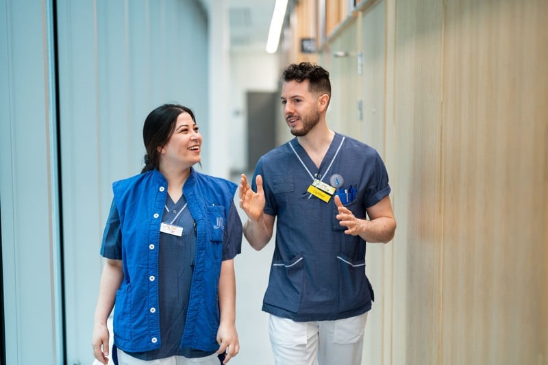 Två personer i blå sjukhuskläder går genom en ljus korridor och pratar med varandra. De ser glada ut och engagerade i samtalet.