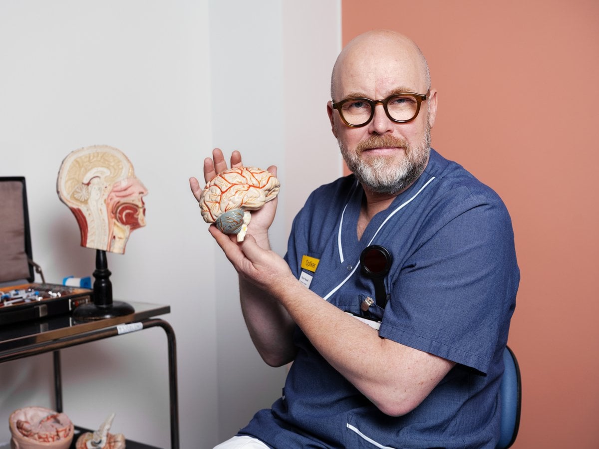 En man med glasögon och sjukhuskläder håller i en modell av en hjärna.