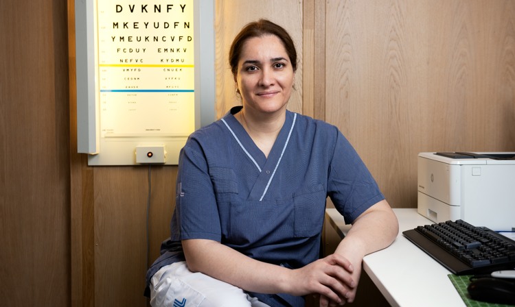 En kvinna i sjukhuskläder sitter vid ett skrivbord framför en syntavla.