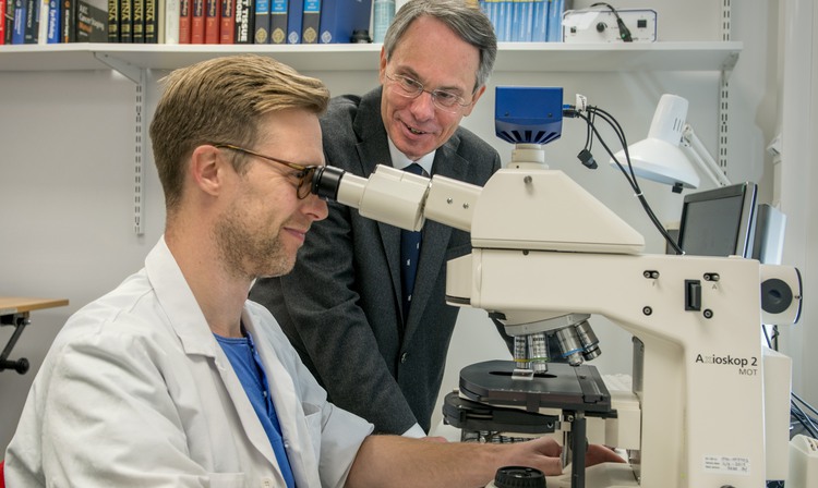 A man in a lab coat in front of a microscope. A man in a suit standing by.