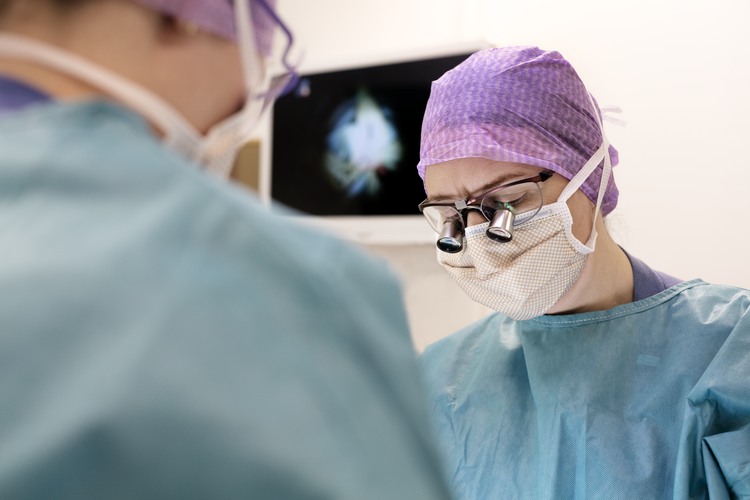 Kvinna i operationsrock, operationsmössa, munskydd och operationsglasögon.