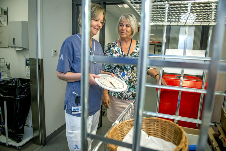 Två kvinnor som ler, den ena i blommig tröja och vit kjol, den andra i sjukvårdskläder. 
