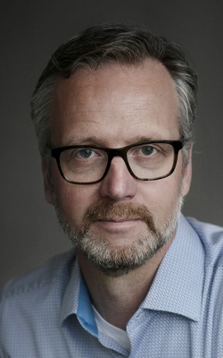 En man med glasögon och skägg i blå skjorta.